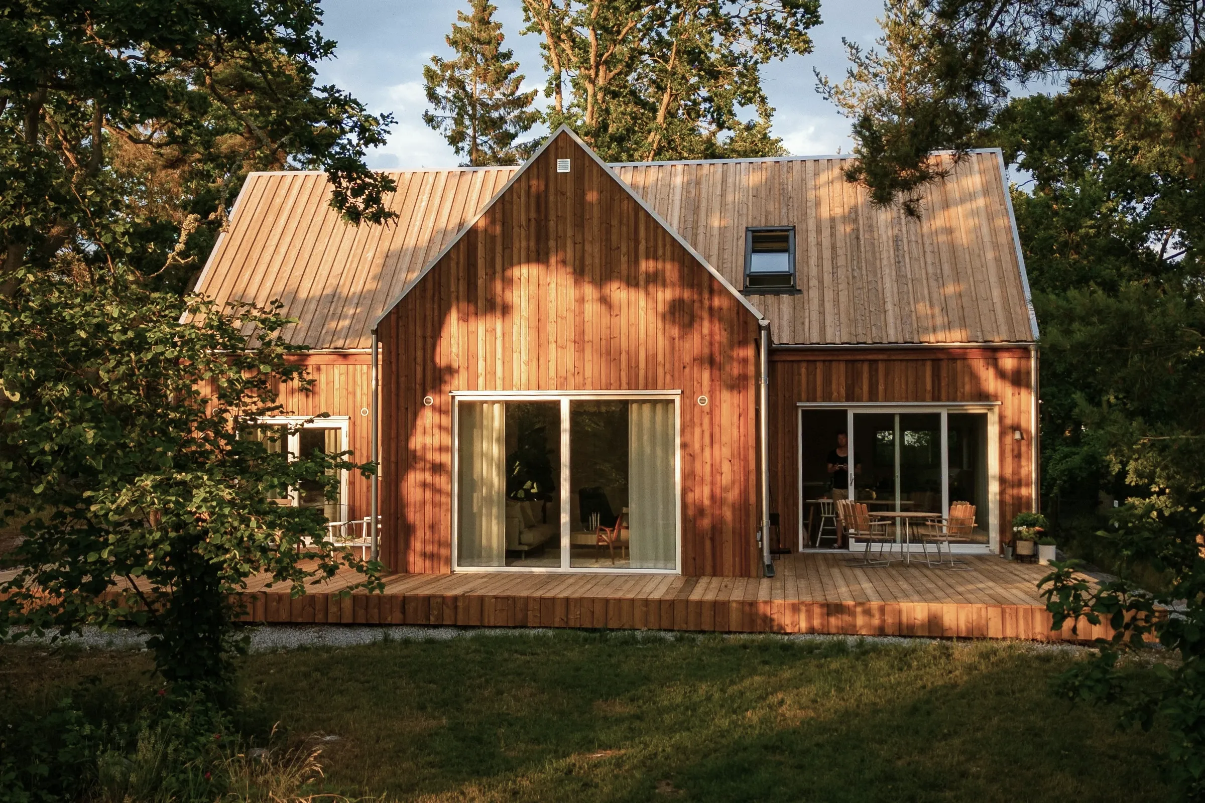 A timber-clad house with a steep metal roof, its sliding glass doors opening onto a wide wooden deck in evening light