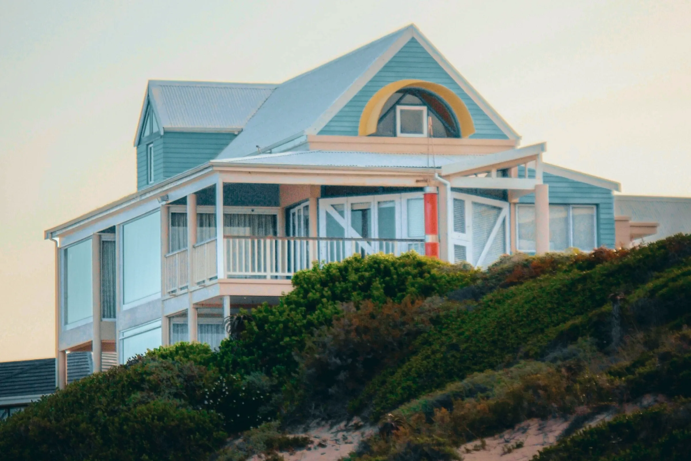 A pale blue weatherboard beach house with a wraparound veranda, standing above a scrub-covered dune at sunset