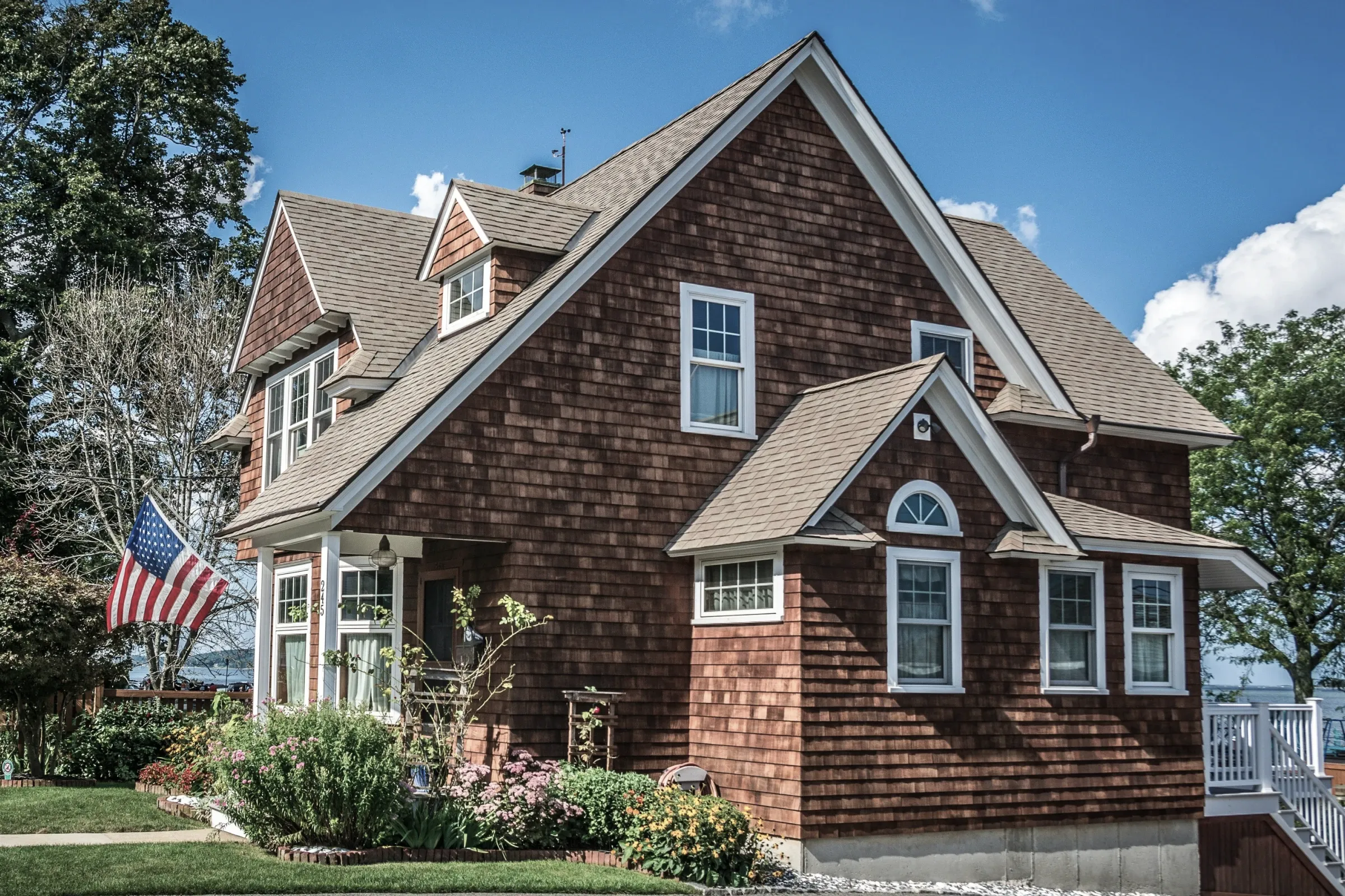 A cedar-shingled house with white-trimmed dormer windows and flower beds along its front path, open water behind it